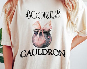May include: Cream-colored t-shirt with the words "BOOKCLUB CAULDRON" in black script. A watercolor illustration of a cauldron with a pink bow is centered on the shirt. The shirt is worn with dark green shorts.