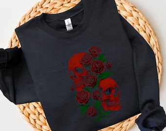 May include: A navy blue sweatshirt featuring a red skull and rose design. The graphic includes two skulls surrounded by red roses and green leaves. The long-sleeved top is displayed on a woven, circular surface.