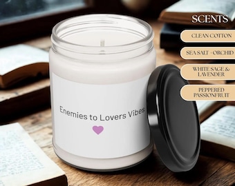May include: A clear glass jar candle with a black lid, labeled "Enemies to Lovers Vibes" with a pink heart. The candle is surrounded by books and lists scents: Clean Cotton, Sea Salt + Orchid, White Sage & Lavender, and Peppered Passionfruit.
