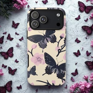 Puede incluir: Una funda de teléfono con un fondo crema con un patrón de mariposas negras y moradas y flores rosas. La funda tiene un módulo de cámara negro y está rodeada de mariposas y flores adicionales.
