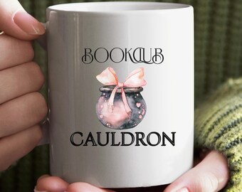 May include: White ceramic mug with the words "BOOKCLUB CAULDRON" in black lettering. A watercolor illustration of a cauldron with a pink bow and pink potion is in the center of the mug. The mug is held by a person.