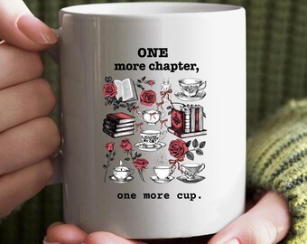 May include: White ceramic mug with the text "ONE more chapter, one more cup." The mug features illustrations of books, roses, teacups, and candles in red and black. The mug is held by a person wearing a green sweater.