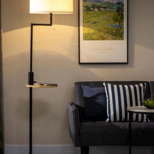 Peut inclure: Un lampadaire noir avec un abat-jour en tissu blanc et une table d'appoint. La lampe a un bras incurvé et une petite étagère pour ranger des objets. La lampe est à côté d'un canapé gris avec des coussins rayés bleus et blancs.