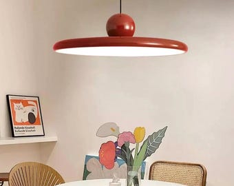 May include: A red pendant light fixture with a round, disc-shaped shade and a spherical top. The light is hanging over a white table with a vase of colourful flowers.