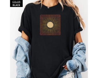 May include: Black t-shirt with a square design. The design features a central circle with radiating lines in gold, orange, and red. The shirt is black. The text "COLOR BLACK" is in the upper left corner.