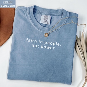 Könnte beinhalten: Ein blaues Jeans-T-Shirt mit dem weißen Text "faith in people, not power". Eine goldene Halskette mit Anhänger liegt über dem Shirt. Das Shirt ist gefaltet und hat ein Comfort Colors-Etikett.