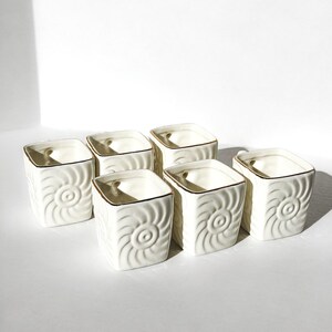 Op de afbeelding: Zes vierkante, crèmekleurige keramische plantenbakken met een spiraalvormig ontwerp en gouden rand. De plantenbakken zijn gerangschikt op een wit oppervlak, waarbij hun decoratieve textuur en elegante ontwerp worden getoond. Ideaal voor kleine planten.