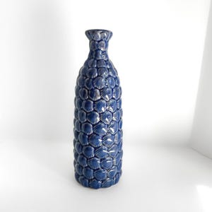 May include: A tall, navy blue ceramic vase with a textured surface. The vase is covered in a repeating pattern of shell-like shapes. The vase has a narrow neck and a slightly flared base. The vase is approximately 12 inches tall.