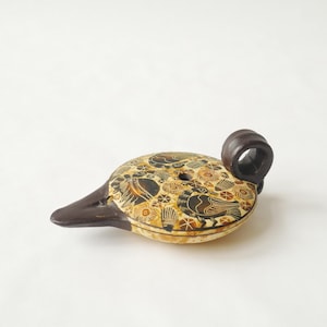 May include: A ceramic oil lamp with a dark brown handle and spout, featuring a cream-colored body decorated with black and brown illustrations of birds, circular patterns, and small geometric shapes across the top surface.