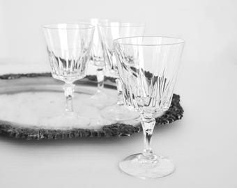 May include: Four clear crystal wine glasses with etched vertical lines, arranged on a silver-colored, ornate tray. The glasses have a classic design with a stemmed base and a flared bowl. The background is a soft, neutral tone.