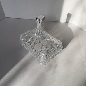 May include: A clear, rectangular crystal candy dish with a lid. The dish features a geometric, cut-glass design and a square handle on the lid. The dish is sitting on a white surface.