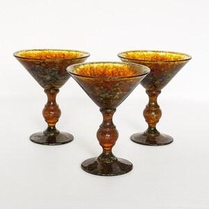 May include: Three vintage-style cocktail glasses. Each glass features a wide, flared bowl and a decorative stem. The glasses have a speckled, amber-colored finish with dark green and brown accents, creating a unique, textured appearance.