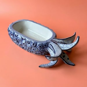 May include: A ceramic bowl with a light blue and white glaze, featuring a textured exterior with repeating circular patterns. The bowl has an elongated oval shape and includes three curved, tentacle-like appendages extending from one end.