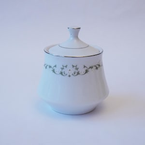 Puede incluir: Un azucarero de porcelana blanca con tapa. El cuenco tiene un ribete plateado y un motivo floral decorativo en verde y blanco en el centro. La tapa tiene un pequeño asa plateada.