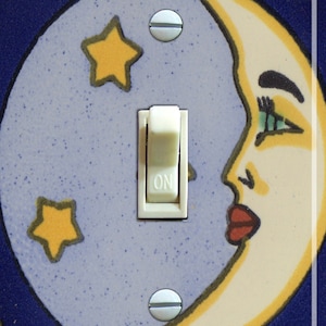 May include: A light switch cover with a crescent moon and stars design. The moon has a face with green eyes and red lips. The switch is labeled "ON".