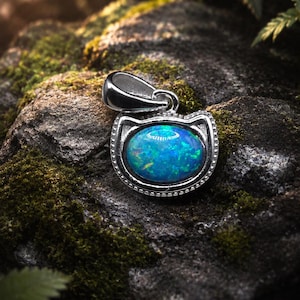 May include: A silver pendant shaped like a cat head featuring a central oval-cut iridescent blue opal gemstone. The metal frame has a beaded edge detail and a polished silver bail at the top for attaching to a chain or necklace cord.