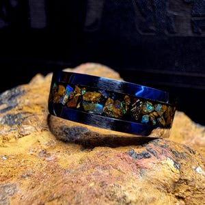 May include: A wide, dark blue ring with a silver band. The center of the ring is filled with a mosaic of gold and iridescent stones. The ring is set against a textured, brown surface.