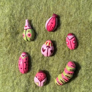 Op de afbeelding: Een verzameling van zeven handgeschilderde, grillige figuren in de vorm van insecten. De figuren hebben verschillende vormen en maten, met roze, groene en rode kleuren. Sommige hebben hart- en stippenpatronen, terwijl andere lijken op rupsen en kevers.