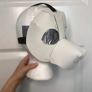 May include: A white animal-style mask crafted from foam or cardstock, featuring a prominent snout and eye openings covered with silver duct tape. The mask is secured to a white styrofoam head form with a black elastic strap across the top and sides.
