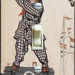 May include: A vintage illustration of a golfer in a plaid suit swinging a golf club. The golfer is standing on a building with a crowd of people watching. The image is printed on a light switch plate with a white toggle switch.