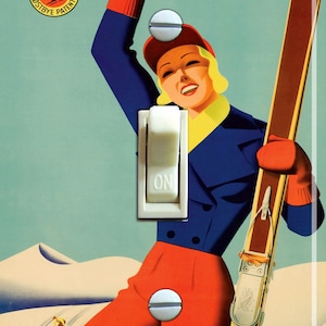 May include: A vintage-style light switch cover featuring a woman in a blue and red ski outfit holding skis. The background is a blue sky with white snow-capped mountains. The text "Ski Tahoe" is at the bottom of the image. The switch cover is labelled "Flexible Flyer" and "Splitkein Ostbye Patent".