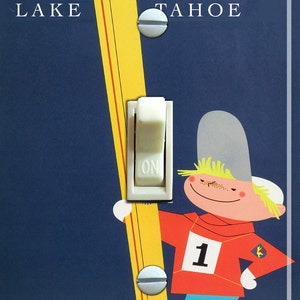 May include: A light switch cover with a blue background and a skier holding a ski. The ski is yellow with a red stripe. The skier is wearing a red and white ski suit with the number "1" on the front. The switch cover says "LAKE TAHOE" at the top.