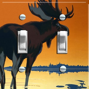 May include: A double light switch plate with a vintage illustration of a moose standing in a lake at sunset. The moose is brown and has large antlers. The background is orange and yellow with a blue lake. The switch plate is white with the word "ON" printed on each switch.