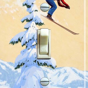 May include: Light switch cover with a retro illustration of a skier in red and blue clothing, mid-air. The background depicts a snowy mountain landscape and a snow-laden evergreen tree. The switch is marked "ON".