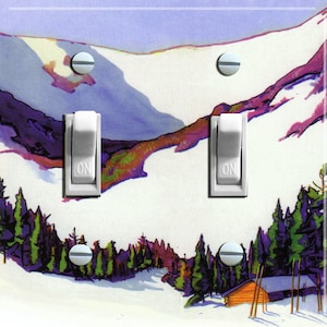 May include: A double light switch plate featuring a winter scene. The image depicts a snowy mountain range with a cabin in the foreground. The switch plate is white with two toggle switches labelled "ON".
