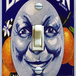May include: A light switch cover featuring a vintage Blue Moon brand orange juice advertisement. The image shows a cartoon moon face with a mischievous grin, surrounded by orange blossoms and oranges. The text on the cover reads "Blue Moon Brand", "Grown on the Selected Groves of H.M. Warren at Highland, Calif."
