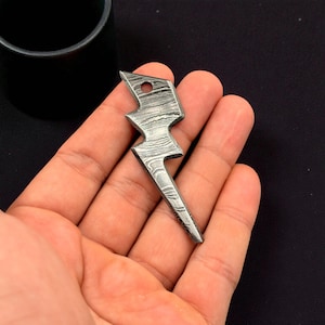 Puede incluir: Un colgante artesanal de metal con forma de rayo con un diseño único. El colgante tiene un pequeño agujero en la parte superior para una cadena o cordón. El metal tiene un aspecto texturizado y en capas, y la forma general es un rayo estilizado.