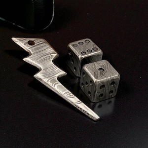 Puede incluir: Un juego de dados de metal y un colgante de rayo con un diseño único. Los dados son cuadrados con esquinas redondeadas y marcas de puntos visibles. El colgante tiene forma de rayo con un agujero en la parte superior.