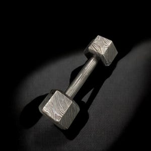 Puede incluir: Una mancuerna de metal estampada con un peso cuadrado en cada extremo, conectada por un mango cilíndrico. La mancuerna es plateada y tiene un patrón similar a la veta de la madera. El fondo es negro.