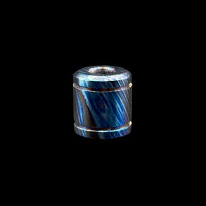 Puede incluir: Un objeto cilíndrico con un patrón arremolinado azul y negro. La parte superior tiene un pequeño agujero centrado. El objeto tiene un brillo metálico y está sobre un fondo negro. El diseño se asemeja a la veta de la madera.