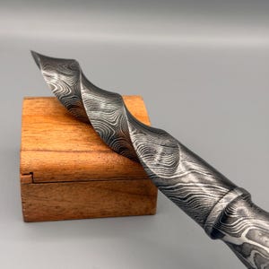 Puede incluir: Primer plano de un cuchillo de acero de Damasco con mango retorcido. La hoja tiene un patrón ondulado y el mango es texturizado. El cuchillo descansa sobre una pequeña caja de madera.