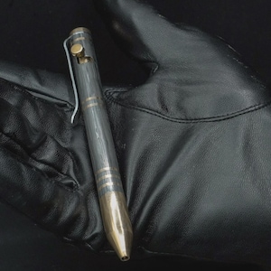 Puede incluir: Primer plano de un bolígrafo con un diseño gris oscuro y dorado. El bolígrafo tiene un cuerpo texturizado con bandas doradas y una punta dorada. Un clip plateado está sujeto al bolígrafo. El bolígrafo descansa sobre una superficie de cuero negro.