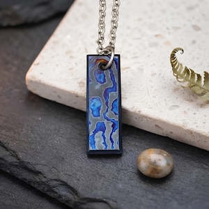 May include: A rectangular pendant necklace featuring a metallic surface with an abstract pattern of vibrant blue, purple, and grey organic shapes. The pendant is suspended from a silver-toned metal chain link, showcasing a polished dark frame.