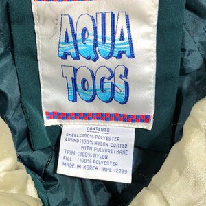 May include: Close-up of a tag from an Aqua Togs jacket. The tag features the brand name in blue and white, with a red and blue checkered border. The tag also lists the materials: polyester, nylon, and polyurethane.
