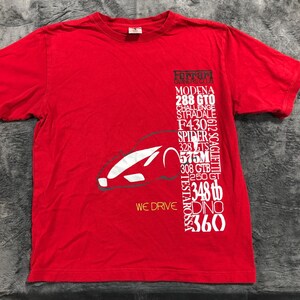 May include: Red t-shirt featuring a white car graphic and the text "WE DRIVE". The shirt also lists various Ferrari models, including Modena, 288 GTO, and Testarossa. The shirt is made of a soft material.