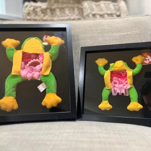 May include: Two framed plush frog toys with green bodies and yellow extremities. Each frog features a yellow vest and an open midsection revealing a collection of pink and white beads, charms, and fabric ruffles. Both items include a red Ty heart tag.