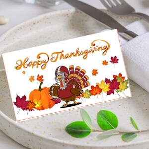 May include: Thanksgiving greeting card featuring a cartoon turkey wearing a football helmet and holding a football. The card has the words "Happy Thanksgiving" in orange script, with pumpkins and autumn leaves. The card is on a white plate with silverware and a napkin.