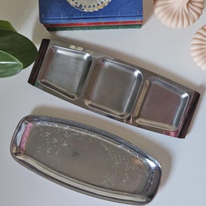 May include: Two vintage serving trays. One is a rectangular tray with a floral design. The other is a stainless steel tray with three square compartments and dark wood accents on each end. The trays are on a white surface.