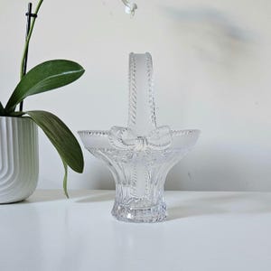 May include: A clear glass basket with a handle and a bow design. The basket has a flared shape and a textured handle. The bow is located on the front of the basket. The basket is sitting on a white surface.