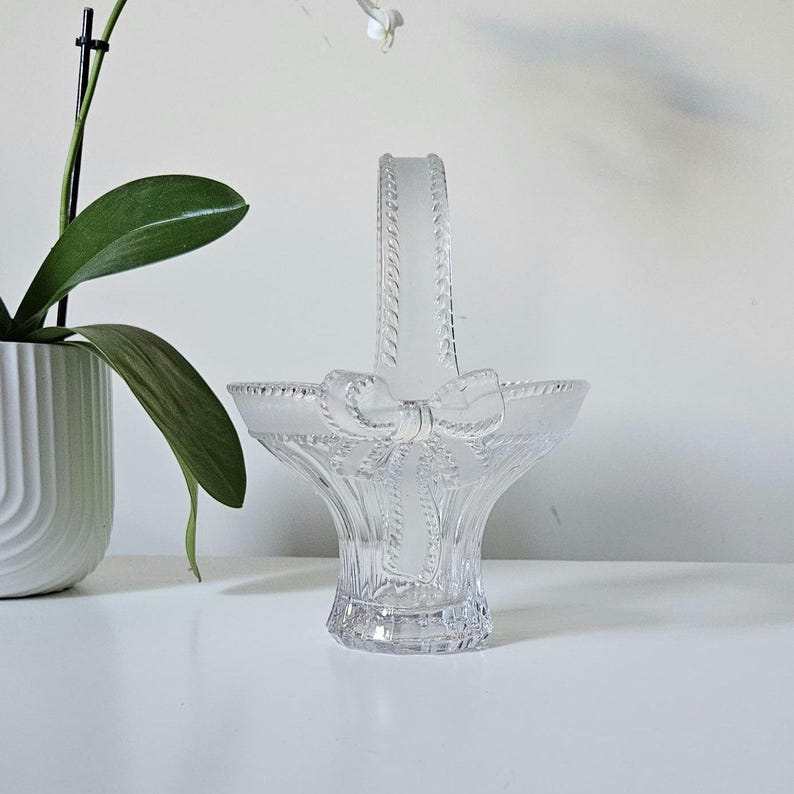 May include: A clear glass basket with a handle and a bow design. The basket has a flared shape and a textured handle. The bow is located on the front of the basket. The basket is sitting on a white surface.