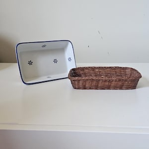 May include: A white rectangular ceramic baking dish with blue floral accents and a dark blue rim sits beside a brown woven basket. The basket is rectangular with rounded edges.