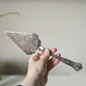 May include: Silver cake server with an intricate floral and scrollwork design. The server has a serrated edge and a decorative handle. A monogram is visible on the blade.