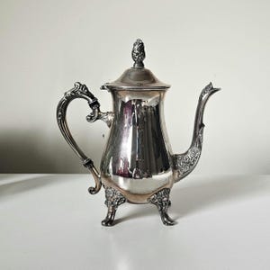 May include: A silver antique coffee pot with decorative features. The pot has a curved handle, a decorative spout, and stands on three small feet. The lid is topped with a decorative finial. The pot is highly reflective, reflecting the surroundings.