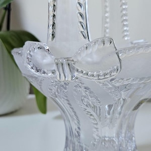 May include: Clear glass basket with a handle and a bow-shaped accent. The basket features a textured design and a frosted finish, creating a delicate and elegant appearance. The handle is adorned with a beaded pattern.