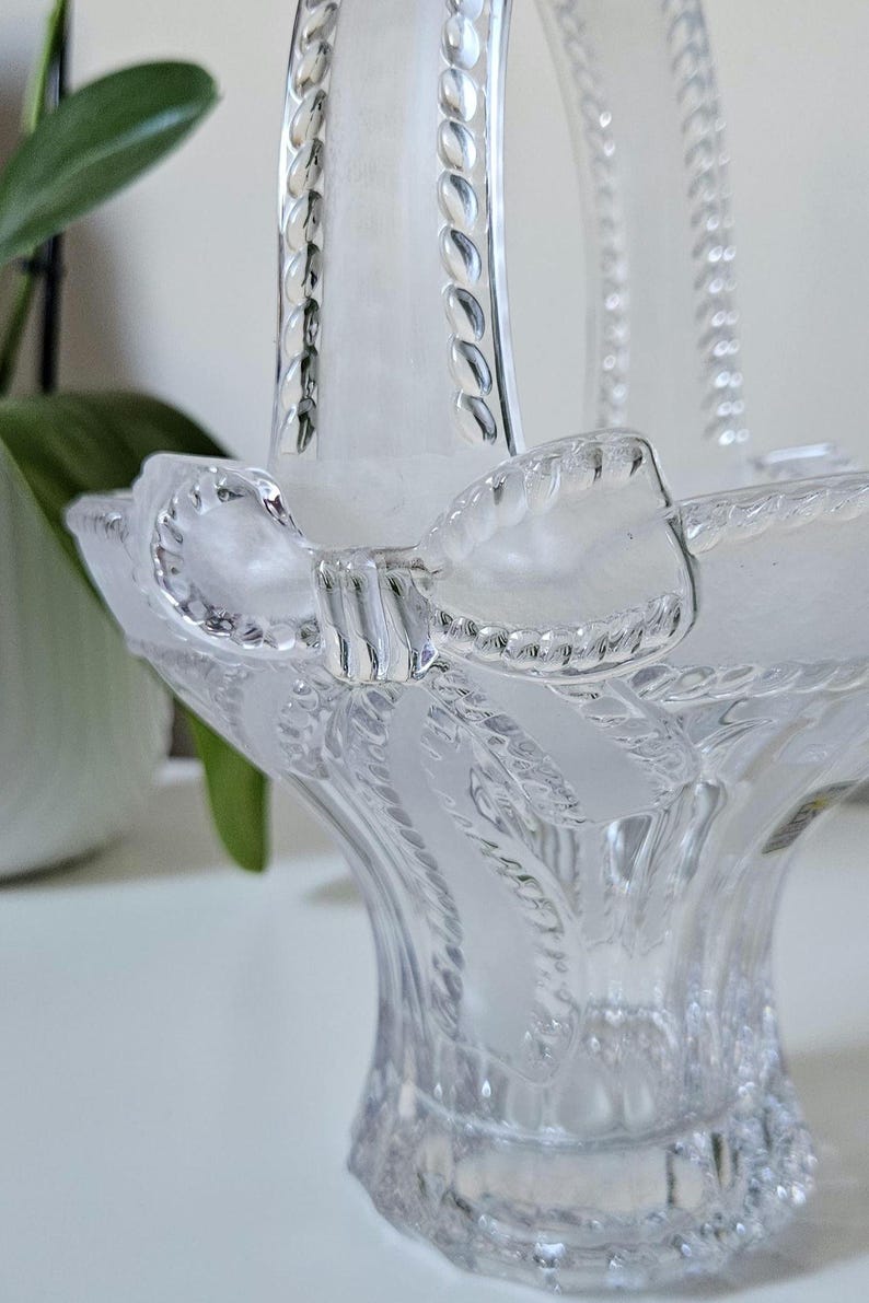 May include: Clear glass basket with a handle and a bow-shaped accent. The basket features a textured design and a frosted finish, creating a delicate and elegant appearance. The handle is adorned with a beaded pattern.
