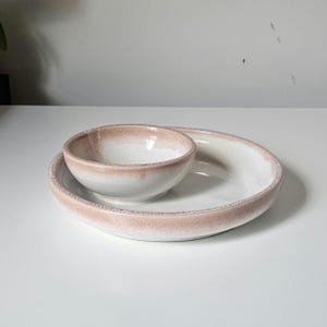 May include: A white ceramic serving dish set with a small bowl nested on a larger, oval-shaped plate. The rims of both pieces have a textured, light brown glaze. The set is designed for serving appetizers or small portions.